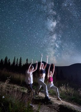 Yıldızlı gecede yoga yapan üç kız. Sportif kızlar samanyolu manzaralı jimnastik yapıyor. Yoga egzersizleri dağlarda.