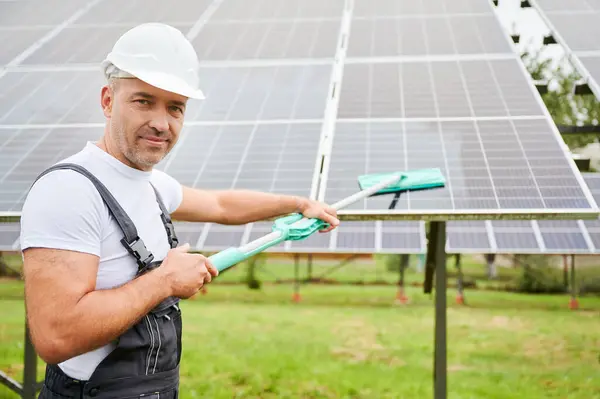 Man cleaning solar PV panel using mop. Professional worker doing good job at cleaning solar panel from dust. Man holding mop and smiling straight to camera.