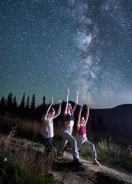 Three girls doing yoga at starry night. Sporty girls doing gymnastics with view of Milky Way. Yoga exercises in mountains.