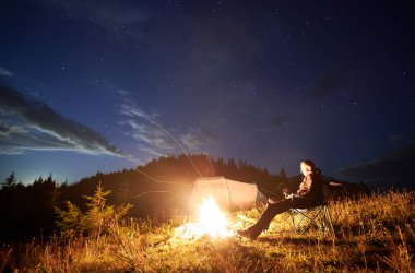 Akşam bulutlu gökyüzünün altındaki dağlarda kamp yapmak. Genç adam kamp ateşinin, turist çadırının ve tepedeki off-road aracının yanında dinleniyor. Gezgin adam oturuyor ve geceleri yıldızlara bakıyor..
