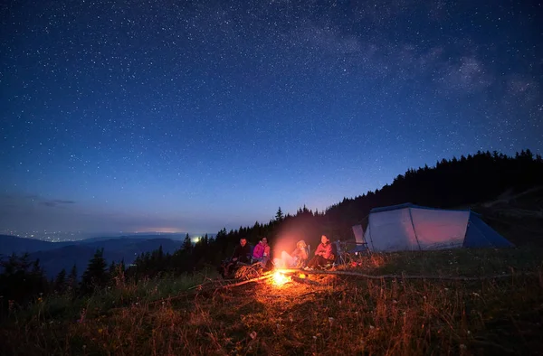 Family camp in mountains. Group of tourists having a rest near campfire on a top of hill, near tourist tent in the evening. Starry night in Carpathian mountains.