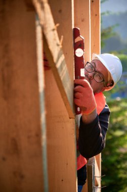 Marangoz ormanın yakınında ahşap çerçeve inşa ediyor. Gözlük takan sakallı adam, ruh seviyesini kullanarak duvarları kontrol ediyor. Koruyucu kask ve turuncu yelek giyiyor..