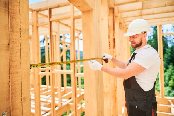 Carpenter building wooden frame house. Man measures the distance with tape measure while dressed in workwear and a helmet. Selective focus.