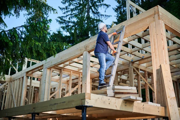 Carpenter constructing wooden frame house near the forest. Bearded man with glasses climbing ladder to the second floor of the two-story building. The concept of modern ecological construction.
