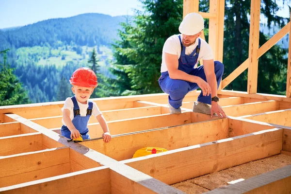 Father with toddler son constructing wooden frame house. Man instructing his son on measurement of distance using tape measure on construction site, wearing helmets and blue overalls on sunny day.
