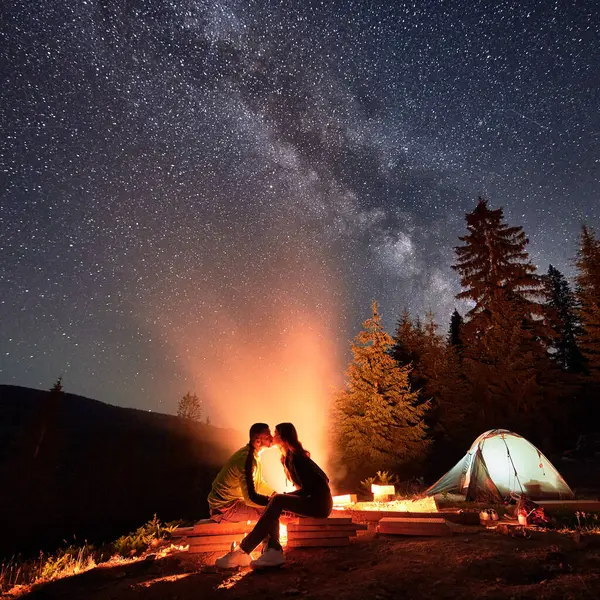 Nocturnal camping adventure amidst mountains, basking in starry splendor. Man and woman kissing near campfire and tent pitched by lush forest, under the captivating constellations of Chumack Path.