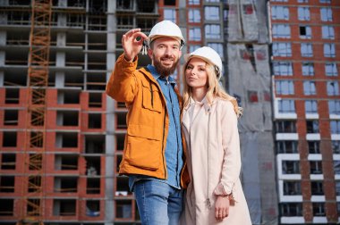 Yeni ev anahtarları olan mutlu çift arka planda inşaat halindeki apartmanlarıyla caddede duruyor. Gülümseyen adam inşaat alanında kadına sarılıyor..