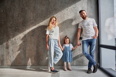 Mutlu bir ebeveyn ve çocuğun portresi. Pencerenin önünde, apartmanın duvarında duruyordu. Erkek ve kadın, yeni evinde, duvarında güneş ışığı gölgesi olan sevimli kızının ellerini tutuyorlar..