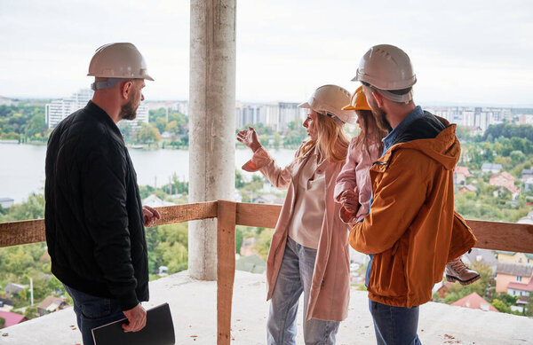 Man and woman with child standing inside apartment building under construction and talking with builder. Family future homeowners discussing building process with construction worker.