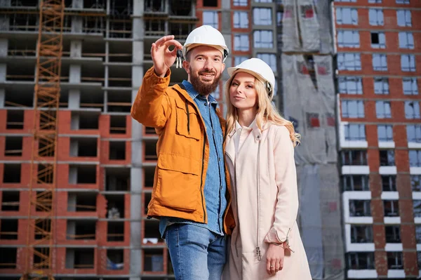Happy couple with new home keys standing on the street with apartment building under construction on background. Smiling man homeowner hugging woman at construction site.