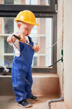 Kid welding electrical wire with electric soldering iron. Child electrician in work overalls and safety helmet installing electrical cables and sockets in apartment under renovation.