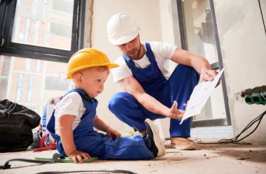 Erkek müteahhit, küçük çocuğa mimari çizimler gösteriyor. Çocuk işçi tulumu ve miğfer giymiş yerde oturuyor ve babamla birlikte apartman planlarını öğreniyor..