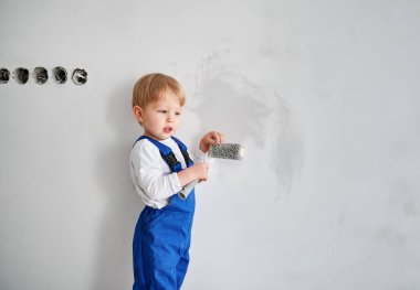 Duvarın yanında duran ve elinde boya silindiri tutan sevimli bir çocuk inşaat işçisi. Güvenlik kasklı ve tulumlu çocuk evde tadilat sırasında boya aleti kullanıyor..