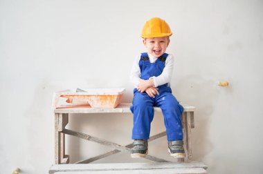 Şirin bir inşaat işçisi, tadilat altındaki beyaz duvarın karşısında, tahta masanın üzerinde oturuyor. Güvenlik kaskı takan ve kameraya bakıp gülen neşeli küçük çocuk..