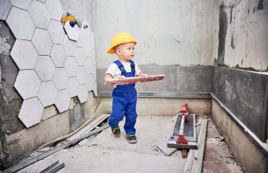Ruh seviyesinde enstrüman tutan erkek bebek inşaat işçisi. Kasklı ve iş tulumlu bir çocuk apartman banyosunda duruyor. Ev yenileme ve çocukluk konsepti.