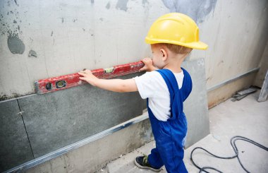 Çocuk inşaat işçisi dairede ruh seviyesindeki aletlerle duvar yüzeyini kontrol ediyor. Çalışan bir çocuk, tadilat sırasında evdeki tamir işleri için duvar hazırlarken aynı zamanda düz bir alet kullanıyor..