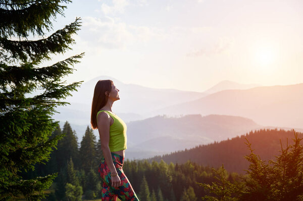 Portrait of young attractive woman posing in the mountains at sunset. Slim, sporty female hiker smiling, admiring landscape, wearing sportswear. Concept of traveling and hiking.