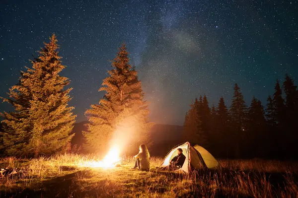 Night camping in mountains under starry sky. Two people, couple having a rest near burning campfire, enjoying beautiful sky full of stars. Concept of tourism and healthy living.