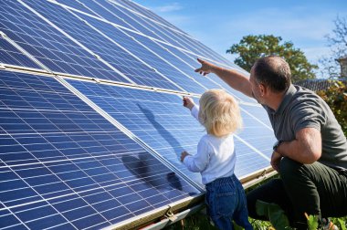 Man showing his small kid solar panels during sunny day. Father presentng to son modern energy resource. Back view of father and his child watching on solar station. clipart