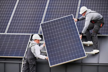 İnşaatçılar evin çatısına güneş paneli inşa ediyorlar. Dışarıda fotovoltaik güneş modülü taşıyan miğferli işçiler. Alternatif ve yenilenebilir enerji kavramı.