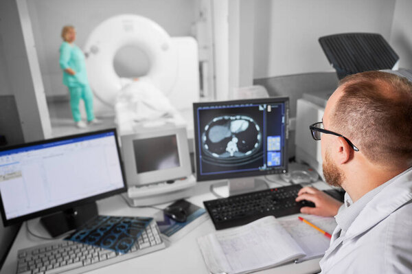 Medical computed tomography or MRI scanner. Back view of male doctor sitting at computer, nurse making MRI on blurred background. Specialists working in clinic. Concept of modern diagnostics.