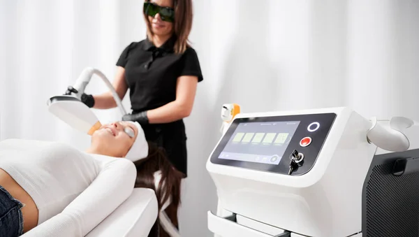 Selective focus of diode laser device cosmetology equipment with cosmetologist and patient on blurred background. Beautician performing laser skincare treatment in beauty clinic.