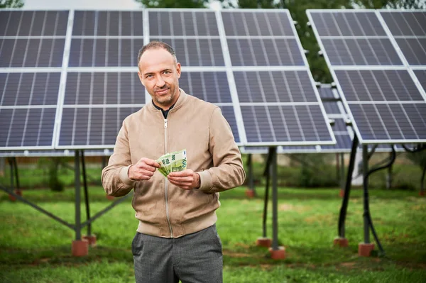 Young male counting money he received by investing in alternative resources. Happy man smiling straight to camera. Man holding euros on background of solar panel.
