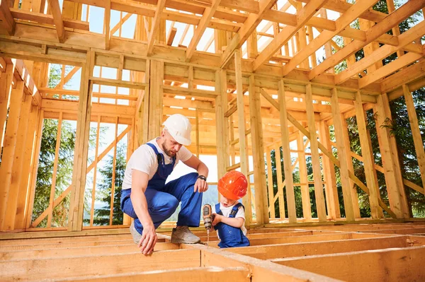 Father with toddler son constructing wooden frame house. Male builder coaching his son on how to operate an electric screwdriver on construction site on sunny day. Carpentry and family concept.