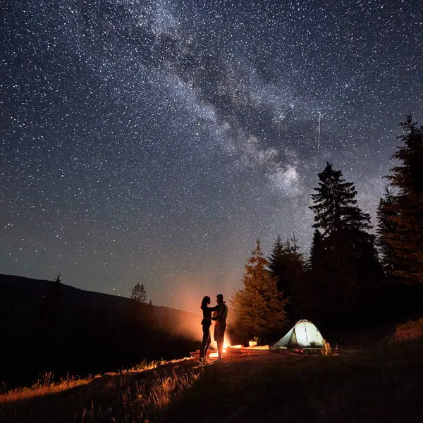 Night camping immersed in starry spectacle. Silhouette of man and woman, avid adventurers, find tranquility by the glowing embers of their campfire, beside their tent nestled amid the pristine forest.