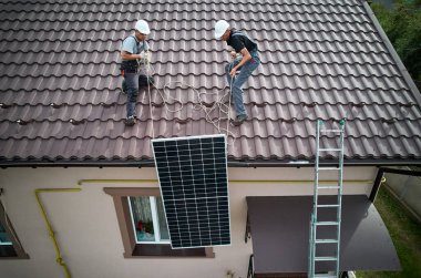 Erkek işçiler fotovoltaik güneş modüllerini evin çatısına kaldırıyorlar. Miğferdeki mühendisler dışarıda güneş paneli sistemi kuruyorlar. Alternatif ve yenilenebilir enerji kavramı. Hava görünümü.