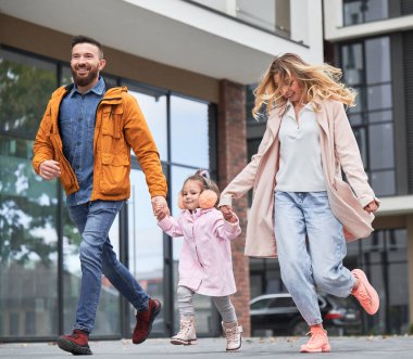 Full length of man and woman holding hands of little girl while walking down the street in new urban district. Happy loving family with child enjoying stroll outdoors near residential building.