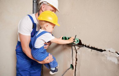 Erkek elektrikçi çocuk duvara elektrik prizleri yerleştirirken sevimli erkek çocuğu tutuyor. Güvenlik kasklı bir adam ve çocuk, tadilat altındaki apartmanda elektrik kabloları yerleştiriyorlar..