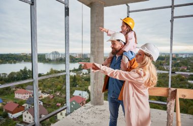 Şantiyede kocasının yanında duran kadın ve kızı tutan adam bir şeyi işaret ediyor. Müstakbel ev sahipleri, inşaat halindeki apartmanı gözetliyor..