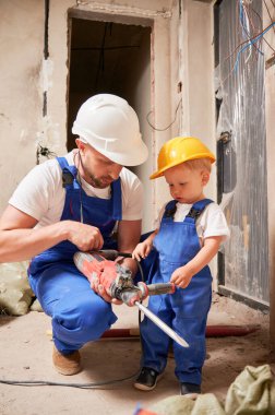 Erkek müteahhit, küçük çocuğa inşaat aletini kullanmayı öğretirken elinde elektrikli çekiç matkabıyla çömeliyor. Tadilat sırasında evde profesyonel sondaj aleti kullanan bir adam ve çocuk..