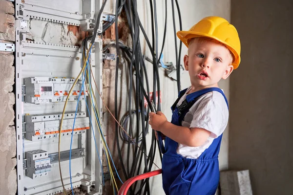 Portrait of child electrician cutting electrical wire with pliers while standing near electrical switch panel. Kid repairing electric wiring cable at home.