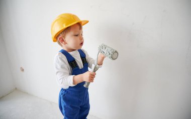 Duvarın yanında duran ve elinde boya silindiri tutan sevimli bir çocuk inşaat işçisi. Güvenlik kaskı takan ve boyama aletleri kullanarak ev yenileme işinde çalışan bir çocuk..