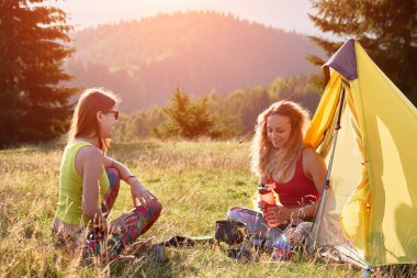 Dağlarda kamp yapan iki kadın turist. Kamp alanında oturan genç kadınlar, mola veriyorlar. Çimenlerde oturan, konuşan, gülümseyen, çay yapan arkadaşlar. Turizm ve yürüyüş kavramı.