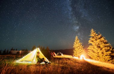 Yıldızlı gökyüzünün altındaki dağlarda gece kampı. Kamp alanındaki çadırın yanında oturan, dinlenen, manzaraya ve kamp ateşine hayran olan bir turist silueti. Yıldızlarla dolu güzel gece gökyüzünün tadını çıkaran kadın..