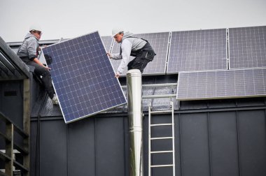 Evin çatısına güneş paneli kuran adamlar. Kasklı elektrikçiler fotovoltaik güneş modülünü açık havada halatların yardımıyla kaldırıyorlar. Alternatif ve yenilenebilir enerji kavramı.