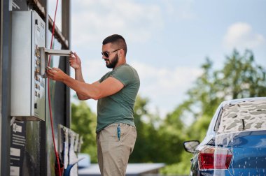 Otomobil yıkamada araba temizleme parasını ödeyen genç adam mavi otomobilin kesilmiş görüntüsü ve onu aktif köpükle yıkayacak adam. Güneşli bir günde arabanın yanında bulaşık için para ödeyen erkek..