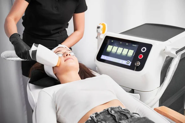 Female patient in safety glasses lying near cosmetology equipment and having face lifting procedure. Doctor cosmetologist using laser scanner device while performing facial treatment in beauty salon.