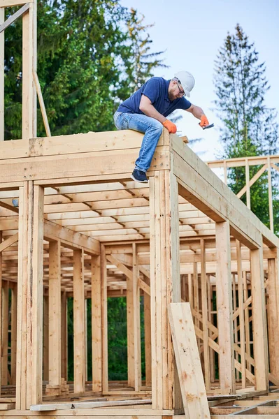 Woodworker constructing wooden, two-story framed house near forest. Bearded man wearing glasses nailing nail with protective helmet on. The concept of modern ecological construction.