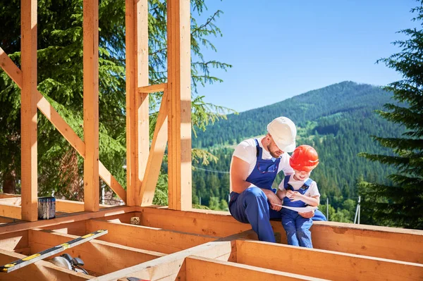 Dad with toddler son constructing wooden frame house. Male builder showing his son the construction plan, wearing helmets and blue overalls on sunny day. Carpentry and family concept.