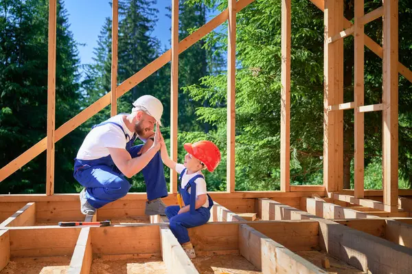 Father with toddler son constructing wooden frame house. Male builder giving high five to kid on construction site, wearing helmets and blue overalls on sunny day. Carpentry and family concept.