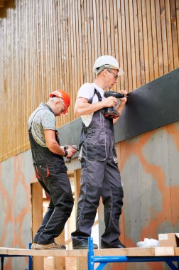 Marangozlar ahşap çerçeveli ev inşa ediyorlar. Çimento parçacık tahtalarıyla evin ön cephesini kaplayan iki erkek işçi onları tornavidalarla bağlıyor. Modern çevre dostu yapı kavramı.