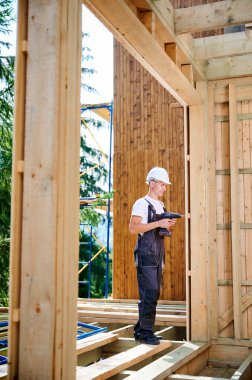 Marangoz ahşap çerçeveli ev inşa ediyor. Evin ön cephesini kaplayan, tornavida kullanan, iş tulumu ve kask giyen bir adam. Modern çevre dostu yapı kavramı.