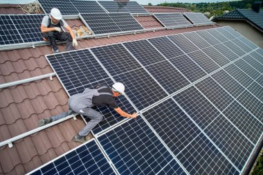 Erkek teknisyen fotovoltaik güneş modüllerini evin çatısına monte ediyor. Kaskın içine monte edilmiş güneş paneli sistemi açık havada kuruluyor. Alternatif ve yenilenebilir enerji kavramı. Hava görünümü.