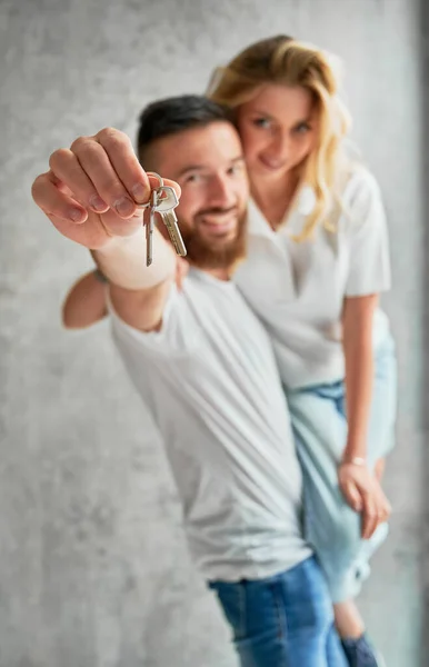 Cheerful man holding wife and smiling while showing apartment keys. Happy couple real estate house buyers demonstrating keys from new home. Focus on male hand with keys.