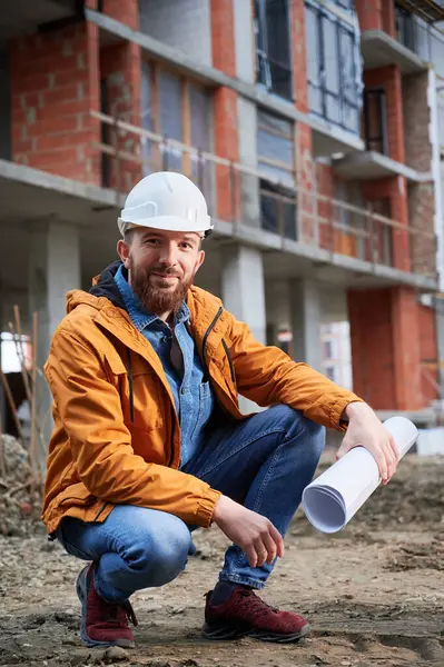 Cheerful man in protective work helmet holding architectural building plan and smiling while crouching down at construction site. Joyful male builder posing outside building under construction.
