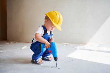 Çocuk inşaat işçisi oyuncak elektrikli matkapla zemini delerken apartmanın duvarının yanında çömelmiş duruyor. Güvenlik kasklı çocuk tadilat sırasında evde elektrikli matkapla çalışıyor..